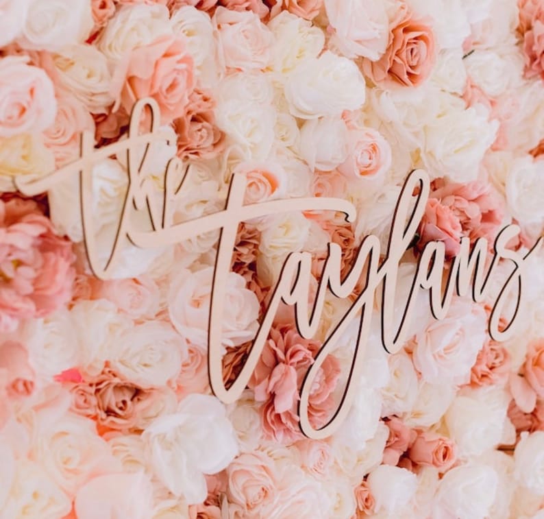 May include: A wooden sign with the name "Taylans" in cursive script is displayed against a backdrop of artificial flowers. The flowers are in shades of white, pink, and peach, creating a soft, romantic aesthetic.