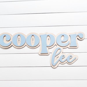 May include: Wooden sign with the name "Cooper Lee" in a layered design. The top layer of the letters is light blue, with a natural wood-colored base. The sign is mounted on a white, horizontal-slat background.
