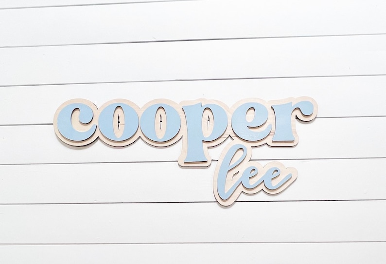May include: Wooden sign with the name "Cooper Lee" in a layered design. The top layer of the letters is light blue, with a natural wood-colored base. The sign is mounted on a white, horizontal-slat background.