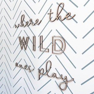 May include: Wooden wall art spelling out "where the wild ones play" in a cursive font. The letters are light brown and mounted on a white wall with a gray chevron pattern.