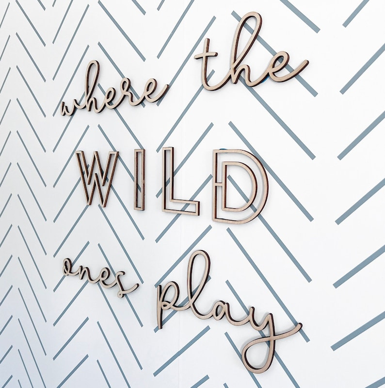 May include: Wooden wall art spelling out "where the wild ones play" in a cursive font. The letters are light brown and mounted on a white wall with a gray chevron pattern.