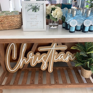 May include: A wooden sign with the name "Christian" in a layered design, displayed on a wooden table. The table also holds mini champagne bottles with blue ribbons, a sign that reads "Babies are sweet please take a treat", and a potted plant.