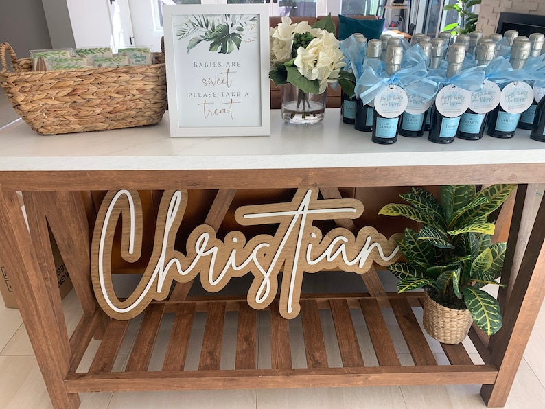 May include: A wooden sign with the name "Christian" in a layered design, displayed on a wooden table. The table also holds mini champagne bottles with blue ribbons, a sign that reads "Babies are sweet please take a treat", and a potted plant.