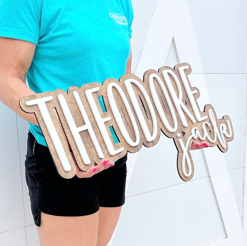May include: A wooden sign with the name "THEODORE jack" in a layered design. The top layer of the letters is white, and the base layer is a light brown wood. The sign is held in front of a white background.