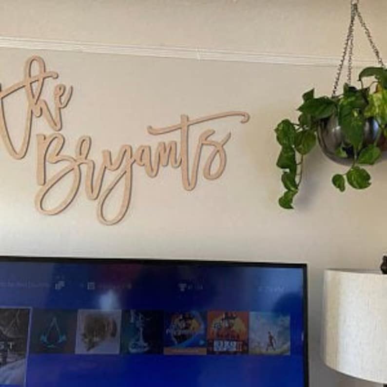 Pu&ograve; includere: Cartello in legno con la scritta "The Bryants" in corsivo, montato su una parete di colore chiaro. Una pianta sospesa in un globo di vetro &egrave; appesa al soffitto. Uno schermo televisivo &egrave; visibile sotto.