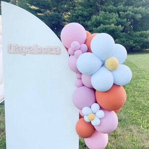 May include: A light blue backdrop with the word "Lilapalooza" in light brown lettering. A balloon garland in shades of pink, orange, and blue is arranged to the right of the backdrop. The garland includes flower-shaped balloon clusters.