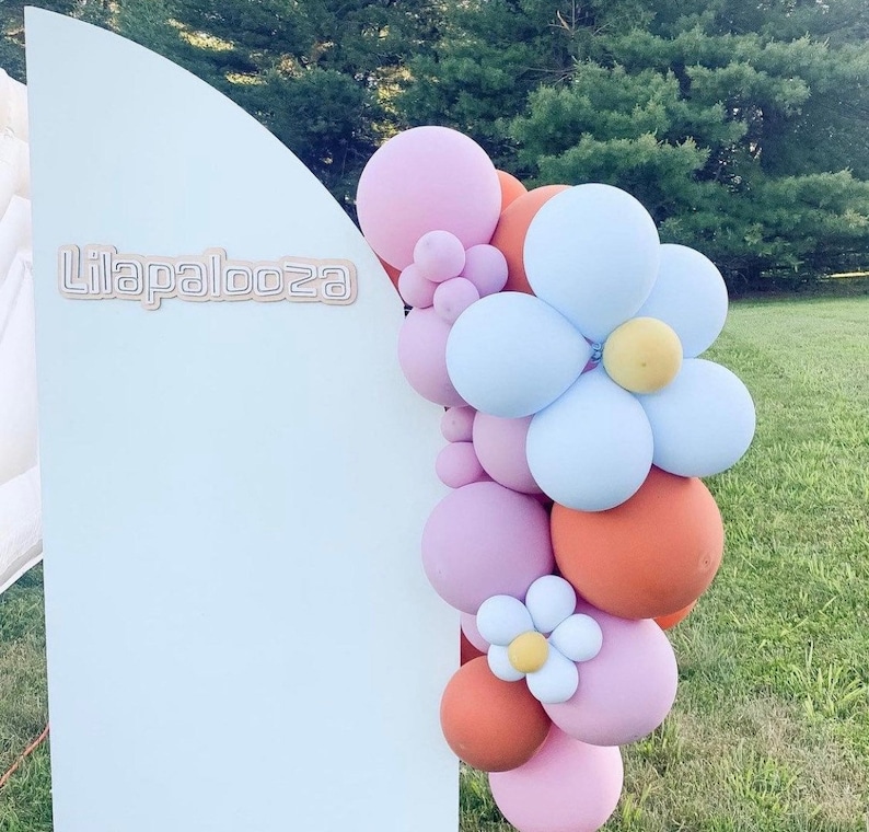 May include: A light blue backdrop with the word "Lilapalooza" in light brown lettering. A balloon garland in shades of pink, orange, and blue is arranged to the right of the backdrop. The garland includes flower-shaped balloon clusters.