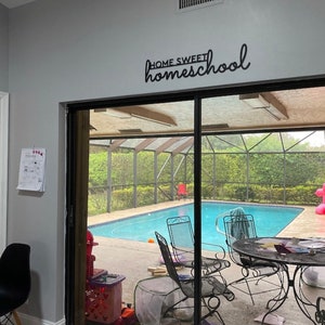 K&ouml;nnte beinhalten: Ein schwarzes Metallschild mit der Aufschrift "HOME SWEET homeschool" ist an einer grauen Wand angebracht. Die Schiebet&uuml;r f&uuml;hrt zu einer Terrasse mit Pool und Gartenm&ouml;beln.