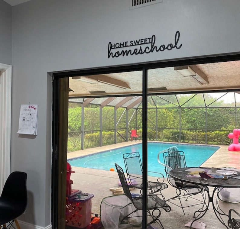 K&ouml;nnte beinhalten: Ein schwarzes Metallschild mit der Aufschrift "HOME SWEET homeschool" ist an einer grauen Wand angebracht. Die Schiebet&uuml;r f&uuml;hrt zu einer Terrasse mit Pool und Gartenm&ouml;beln.