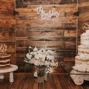 May include: A rustic wedding scene featuring two cakes, floral arrangements, and a wooden backdrop. One cake has a "B" topper, the other a "Mrs. Burke" topper. A vase of white and pink flowers sits in front of the backdrop with the words "the Burkes" in cursive.