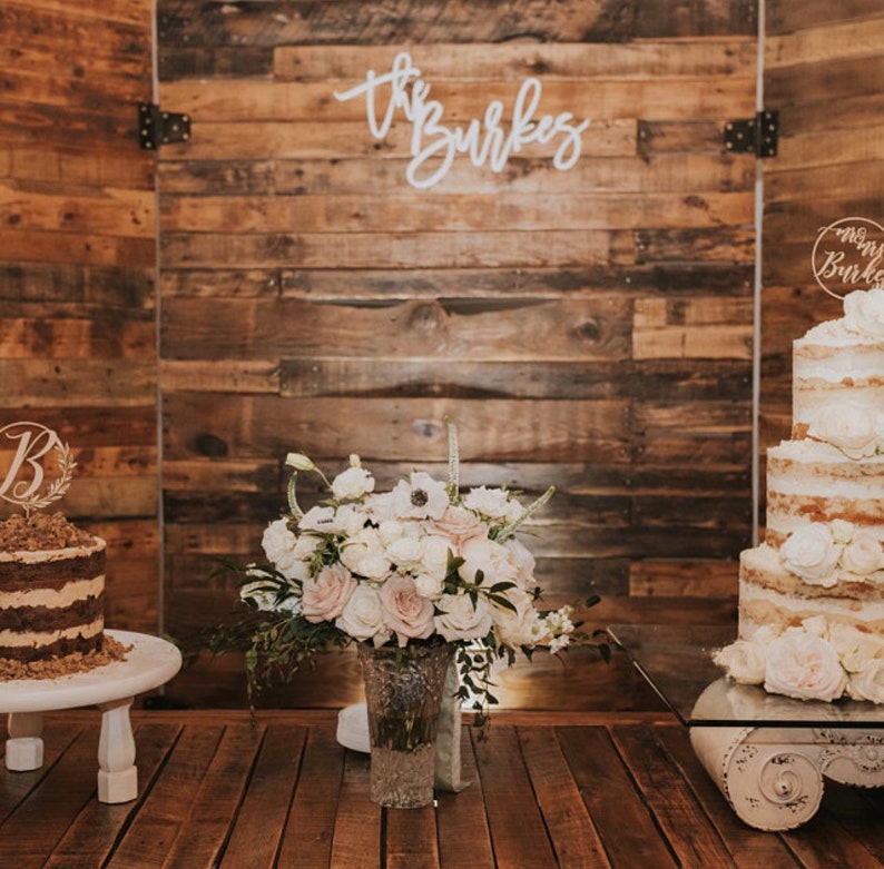 May include: A rustic wedding scene featuring two cakes, floral arrangements, and a wooden backdrop. One cake has a "B" topper, the other a "Mrs. Burke" topper. A vase of white and pink flowers sits in front of the backdrop with the words "the Burkes" in cursive.