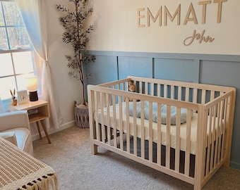 May include: A light wood crib with a mattress and a blue pillow is in a nursery. The wall above the crib has the name "EMMATT John" in wooden letters. A tall plant is in a woven basket.