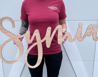 Könnte beinhalten: Holzschild mit dem Namen "Synia" in Schreibschrift. Das Schild besteht aus hellfarbenem Holz und wird vor einem weißen Hintergrund gehalten. Das Schild ist ein dekorativer Artikel.