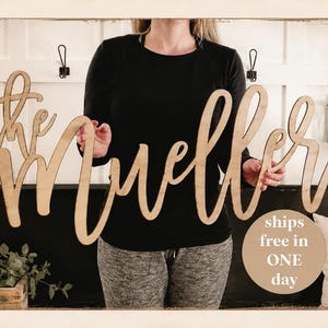 May include: A wooden sign that reads "the Muellers" in a cursive font. The sign is held in front of a white wall with black hooks and a black bench. A pillow with the words "grace upon grace" is visible.