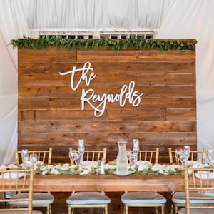 Pu&ograve; includere: Uno sfondo in legno rustico con il testo bianco in corsivo "the Reynolds". Un lungo tavolo di legno &egrave; apparecchiato con sedie dorate, bicchieri e composizioni floreali. Verde e tende bianche completano la scena.