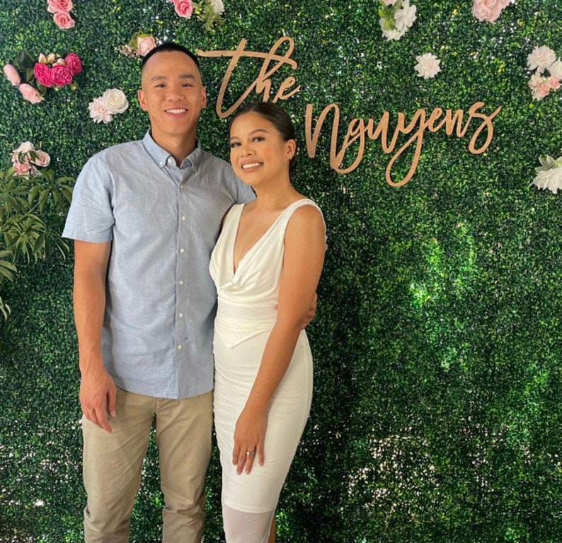 Pu&ograve; includere: Una coppia posa davanti a un muro di siepi verdi decorato con fiori bianchi e rosa. Le parole "The Nguyens" sono visualizzate in caratteri di legno. La donna indossa un vestito bianco e l'uomo una camicia blu e pantaloni color cachi.