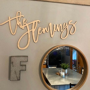 May include: Wooden sign in cursive script that reads "the Hemmings" mounted on a light gray wall. A metal letter "F" and a round mirror with a wooden frame are also on the wall.