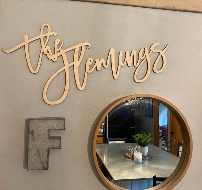 May include: Wooden sign in cursive script that reads "the Hemmings" mounted on a light gray wall. A metal letter "F" and a round mirror with a wooden frame are also on the wall.