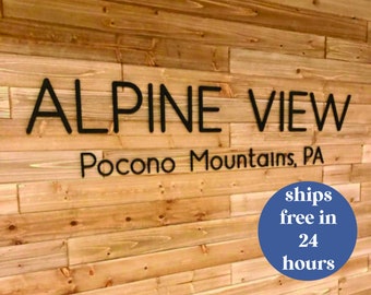 May include: Black lettering spelling "ALPINE VIEW" above "Pocono Mountains, PA" on a light brown wooden wall. The wood planks are arranged horizontally. The text is in a sans-serif font.