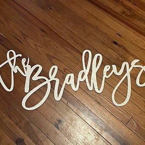 May include: A wooden sign in a cursive font that reads "the Bradleys". The sign is cut from light-colored wood and is displayed on a wooden surface.