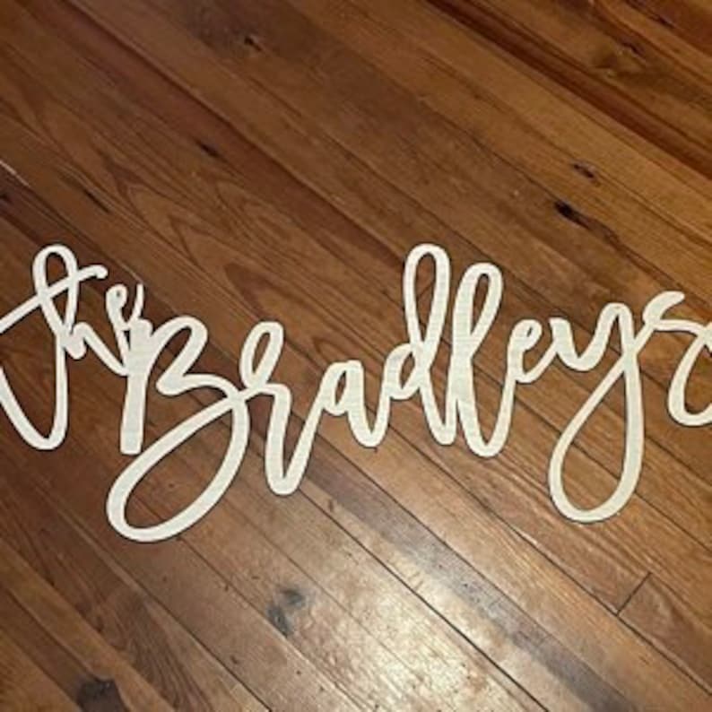 May include: A wooden sign in a cursive font that reads "the Bradleys". The sign is cut from light-colored wood and is displayed on a wooden surface.
