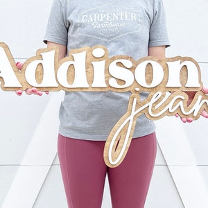 May include: Wooden sign with the name "Addison Jean" in a layered design. The top layer features the name "Addison" in white, while the bottom layer has "jean" in a cursive font. The sign is held in front of a white background.