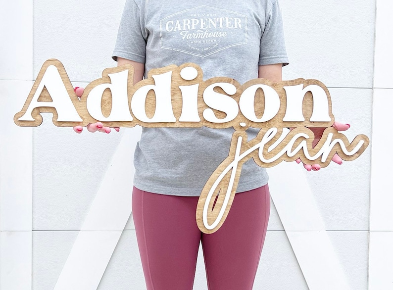 May include: Wooden sign with the name "Addison Jean" in a layered design. The top layer features the name "Addison" in white, while the bottom layer has "jean" in a cursive font. The sign is held in front of a white background.