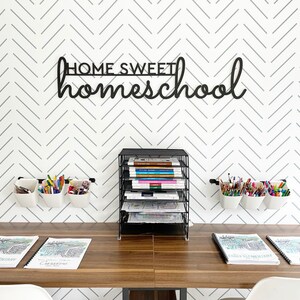 K&ouml;nnte beinhalten: Ein Homeoffice-Setup mit einem Holztisch, einem schwarzen Draht-Organizer und wei&szlig;en Wandbeh&auml;ltern, die mit Stiften und Bleistiften gef&uuml;llt sind. Die Wand hat ein grau-wei&szlig;es Zickzackmuster und ein schwarzes Schild mit der Aufschrift "HOME SWEET homeschool".