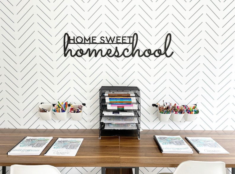 K&ouml;nnte beinhalten: Ein Homeoffice-Setup mit einem Holztisch, einem schwarzen Draht-Organizer und wei&szlig;en Wandbeh&auml;ltern, die mit Stiften und Bleistiften gef&uuml;llt sind. Die Wand hat ein grau-wei&szlig;es Zickzackmuster und ein schwarzes Schild mit der Aufschrift "HOME SWEET homeschool".