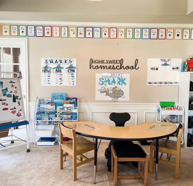 K&ouml;nnte beinhalten: Ein Homeschool-Klassenzimmer mit einem Holztisch und St&uuml;hlen, einer Tafel und einem B&uuml;cherregal voller B&uuml;cher. Die W&auml;nde sind mit Lernpostern geschm&uuml;ckt, darunter ein Alphabetbanner und Lernmaterialien zum Thema Haie.