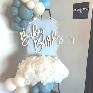 Puede incluir: Decoración de baby shower con un letrero que dice "Baby Burks" en escritura blanca. El letrero está sobre un fondo azul claro, con material blanco en forma de nube y globos azules y blancos. Un letrero que dice "Recreation 3" está en el fondo.
