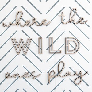 May include: Wooden wall art spelling out "where the wild ones play" in a cursive and block letter font. The letters are light brown and are against a white and gray chevron patterned background.