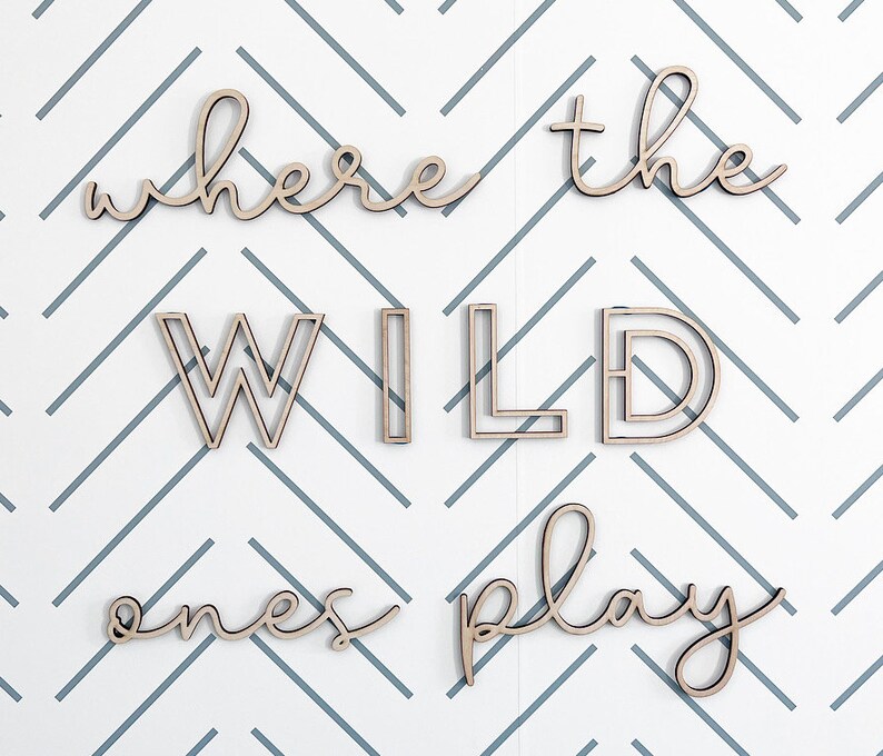 May include: Wooden wall art spelling out "where the wild ones play" in a cursive and block letter font. The letters are light brown and are against a white and gray chevron patterned background.
