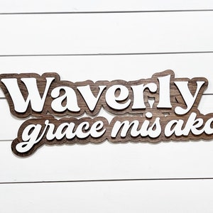 May include: A wooden sign with the name "Waverly grace misako" in a layered design. The top layer features white lettering, while the base layer has a dark wood grain appearance. The sign is mounted on a white, horizontal-slatted background.