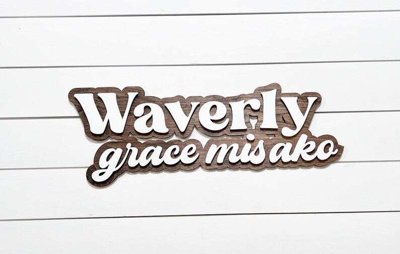 May include: A wooden sign with the name "Waverly grace misako" in a layered design. The top layer features white lettering, while the base layer has a dark wood grain appearance. The sign is mounted on a white, horizontal-slatted background.