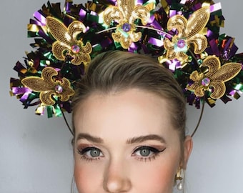Op de afbeelding: Een Mardi Gras-hoofdband met gouden fleur-de-lis-ontwerpen, versierd met pailletten en edelstenen. De hoofdband is versierd met paars, groen en goudkleurig tinsel, wat een feestelijk accessoire creëert.