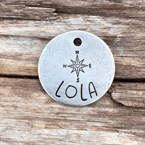 Puede incluir: Chapa redonda de perro de plata con un diseño de brújula y el nombre "LOLA" grabado en ella.
