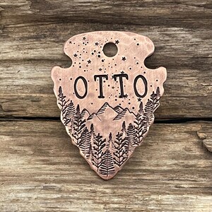 May include: A copper-colored arrowhead-shaped pet tag with a mountain and forest scene engraved on it. The tag has a hole at the top for attaching to a collar. The name "OTTO" is engraved in the center of the tag.
