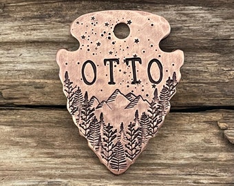 May include: A copper-colored arrowhead-shaped pet tag with a mountain and forest scene engraved on it. The tag has a hole at the top for attaching to a collar. The name "OTTO" is engraved in the center of the tag.