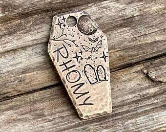 May include: A brass coffin-shaped tag with a hammered finish. The tag is engraved with the name "Rhomy" and a spooky Halloween design featuring bats, stars, a full moon, and tombstones.