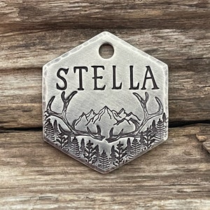 May include: Silver hexagonal pet tag with a mountain and antler design. The tag is engraved with the name "STELLA".