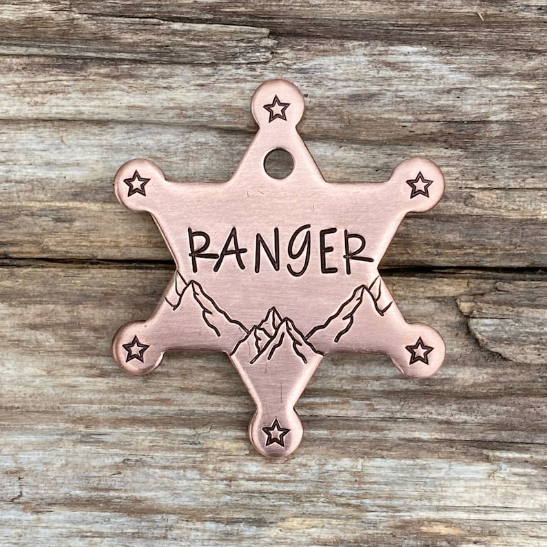 May include: A copper-colored star-shaped dog tag with the word "RANGER" engraved on it. The tag also features a mountain range design.