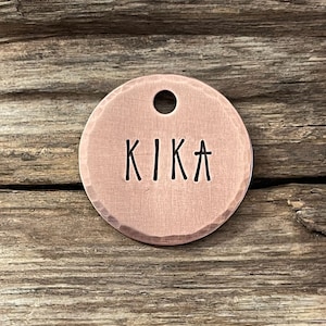 Pode incluir: Uma etiqueta redonda de cobre para cães com acabamento martelado e o nome "KIKA" gravado em preto.