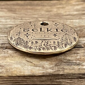 May include: A round brass tag with a forest scene engraved on it. The tag has the word "Selkie" engraved on it, along with stars and mushrooms.