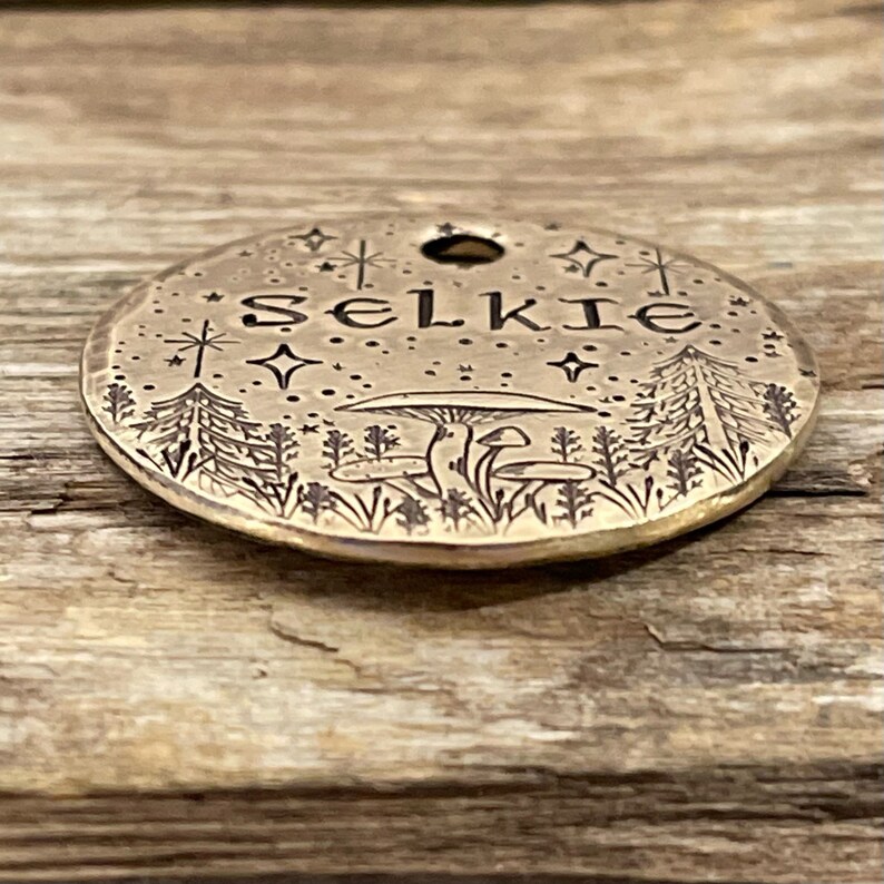 May include: A round brass tag with a forest scene engraved on it. The tag has the word "Selkie" engraved on it, along with stars and mushrooms.