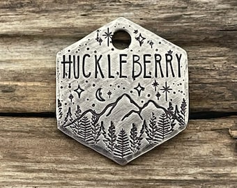 May include: Silver hexagonal dog tag with the word "HUCKLEBERRY" engraved in a cursive font. The tag also features a mountain scene with a crescent moon and stars.