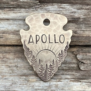 May include: A brass arrowhead-shaped tag with the name "APOLLO" engraved on it. The tag features a sun and trees design.