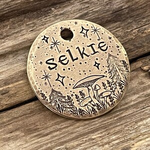 May include: A round brass tag with a hole for a ring. The tag has an etched design of stars, mushrooms, and pine trees. The word "Selkie" is etched in the center.