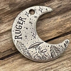 May include: A silver crescent moon charm with the name "RUGER" engraved on it. The charm is decorated with a forest scene including mushrooms, pine trees, and stars.