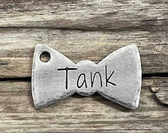May include: A silver bow tie-shaped tag with a hole for a ring. The tag is engraved with the name "Tank".